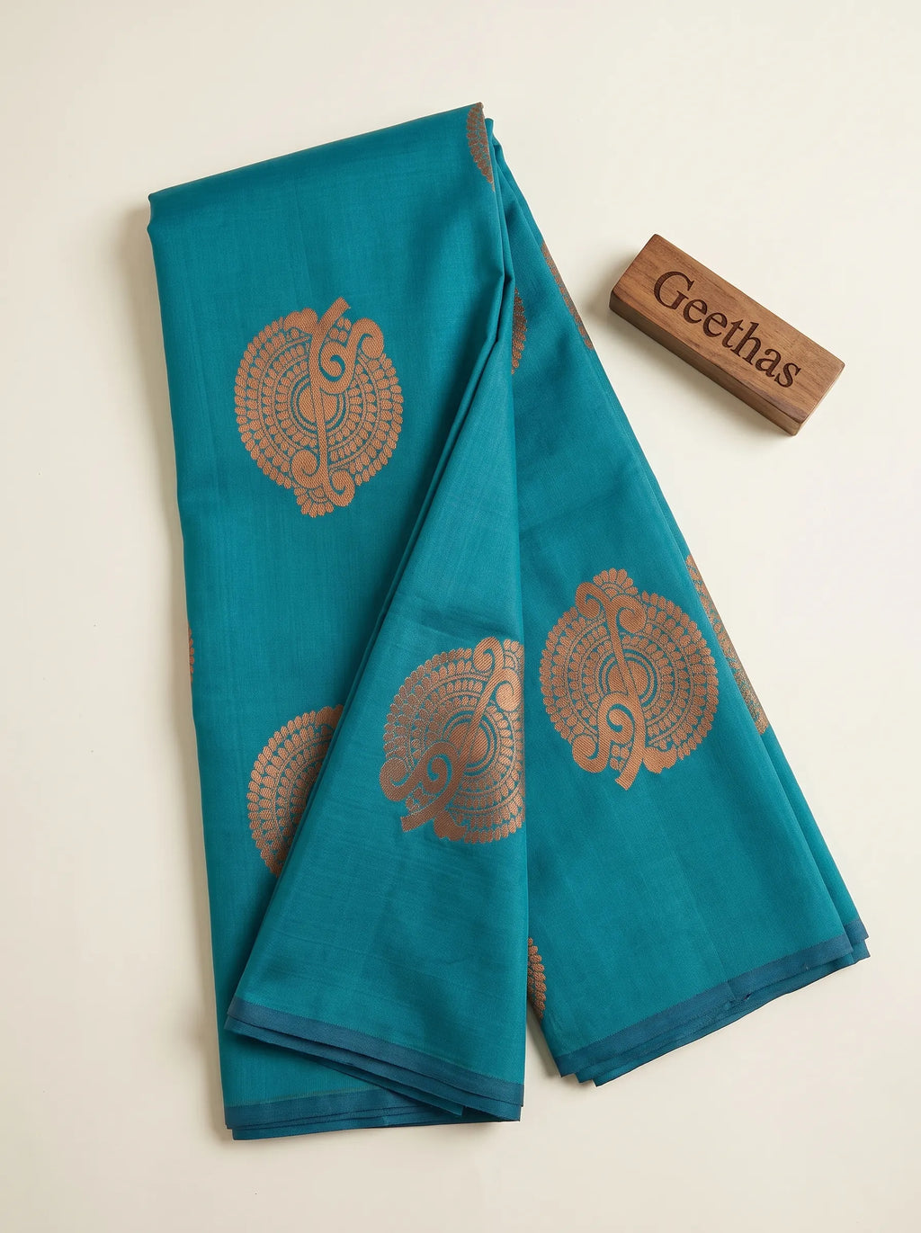 Folded teal blue soft silk saree showing copper-toned woven circular motifs